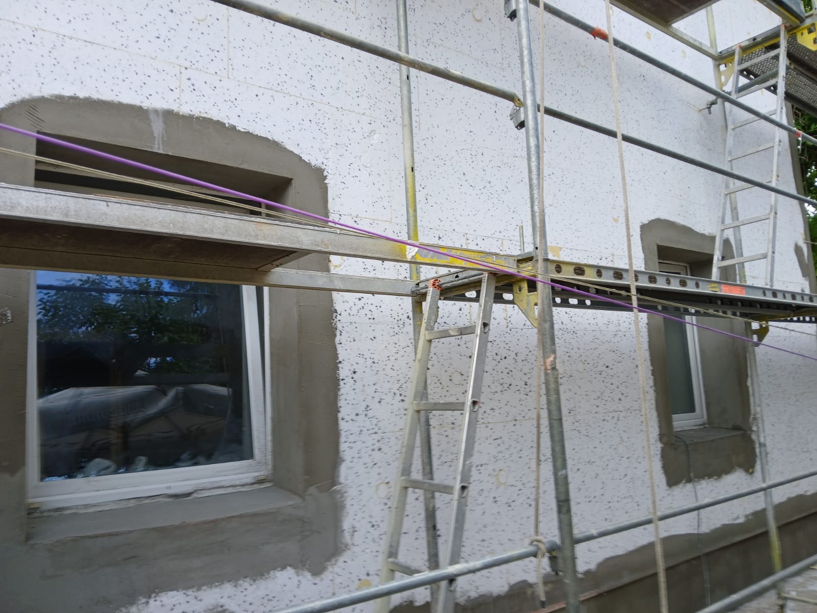 Ocieplanie ściany zewnętrznej budynku styropianem, z widocznymi oknami w trakcie obróbki tynkarskiej, rusztowanie metalowe z drabinką, fioletowe sznurki wyznaczają poziom.