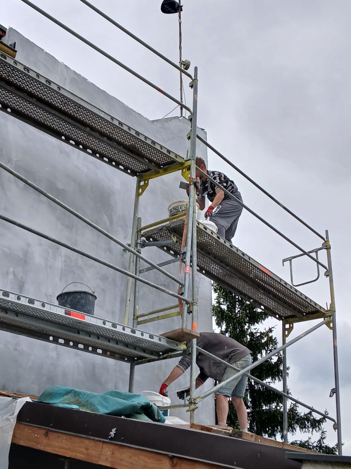 Dwóch pracowników na rusztowaniu, jeden nakłada szpachlą szarą masę na elewację budynku, drugi pracuje poniżej. Widoczny kubeł z masą, narzędzia i zabezpieczenia.