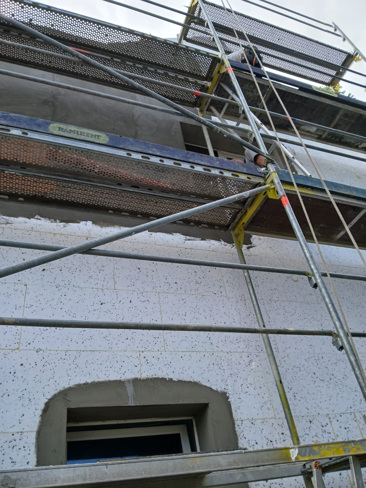 Budynek w trakcie ocieplania, widoczne rusztowanie, ściana z białych bloczków z zarysowanymi otworami okiennymi, fragment otynkowanego otworu okiennego.