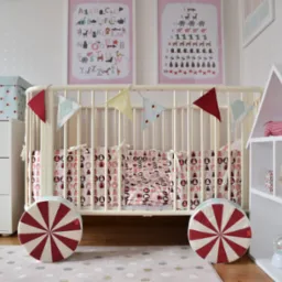 Pokój dziecięcy z białym łóżeczkiem, girlandą z trójkątów, poduszkami w misie, komodą z pudełkiem, regałem w kształcie domku i plakatami edukacyjnymi na ścianie w gwiazdki.