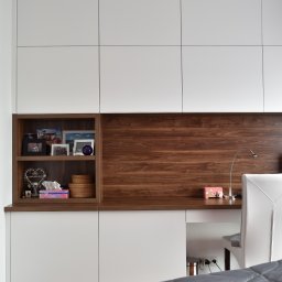 PODLASKA AGENCJA BUDOWLANA MARTA I ADAM BUCZYŃSCY SPÓŁKA CYWILNA - Białe szafy na wymiar z drewnianymi akcentami, półka z dekoracjami, biurko z lampką, widok z perspektywy łóżka z szarą narzutą.