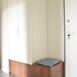 PODLASKA AGENCJA BUDOWLANA MARTA I ADAM BUCZYŃSCY SPÓŁKA CYWILNA - Wysoka biała szafa z drewnianymi dolnymi szafkami i siedziskiem z poduszką we wzory, obok białe drzwi wejściowe z zamkiem i kluczami, heksagonalna szara podłoga.