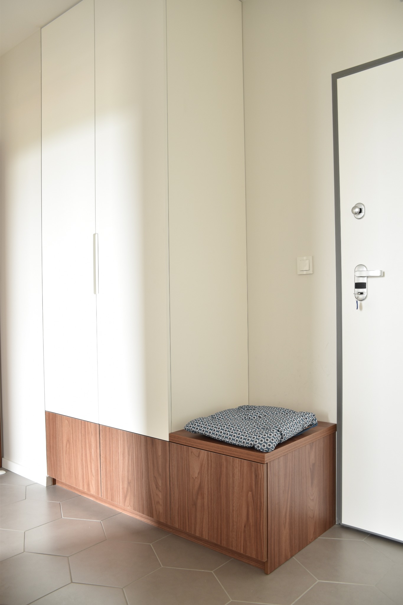 Wysoka biała szafa z drewnianymi dolnymi szafkami i siedziskiem z poduszką we wzory, obok białe drzwi wejściowe z zamkiem i kluczami, heksagonalna szara podłoga.