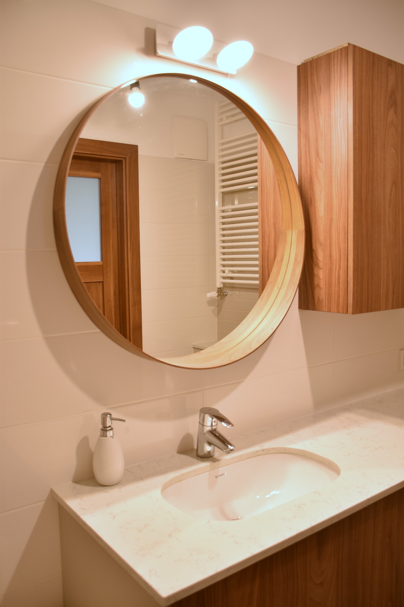 Nowoczesna łazienka z okrągłym lustrem w drewnianej ramie, nablatową umywalką Duravit i szafką z forniru orzechowego, widoczny grzejnik drabinkowy i dozownik mydła.