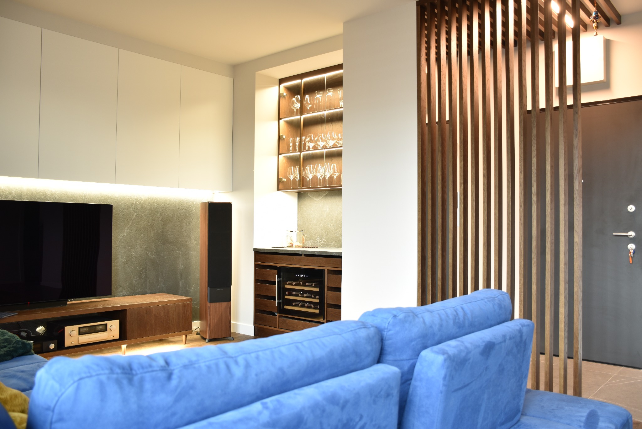 Nowoczesny salon z zabudową ściany TV, barkiem fornirowanym z szafką na wino, ażurową ścianką z lameli i komfortową, niebieską sofą na pierwszym planie.