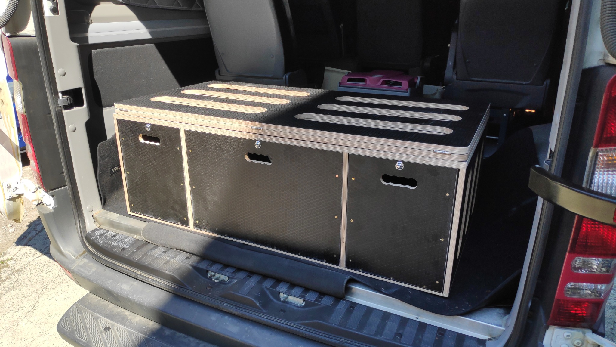 Czarna, drewniana skrzynia z wyciętymi uchwytami i otworami wentylacyjnymi na górze, umieszczona w bagażniku samochodu dostawczego.