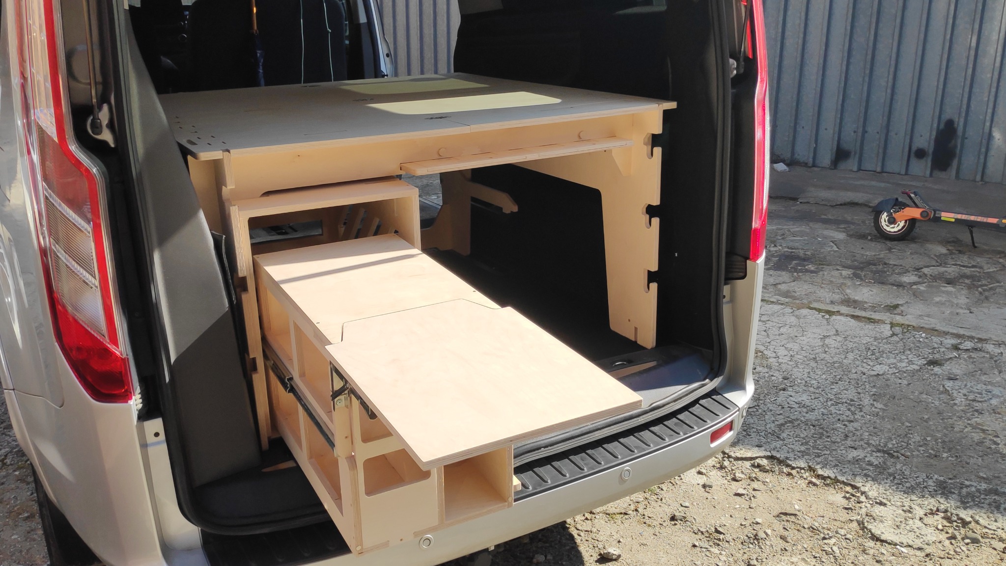 Wnętrze bagażnika samochodu typu van z zabudową modułową z jasnej sklejki, zawierającą wysuwany blat i półki, widoczne elementy mocujące.