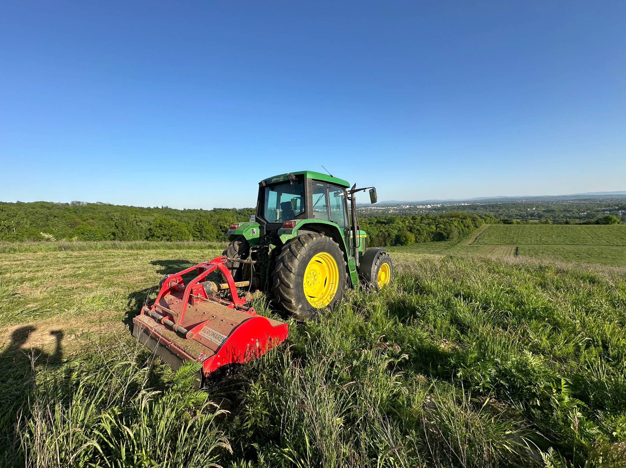 Zielony traktor John Deere z czerwoną kosiarką bijakową Agromarek na łące, widok na oddalone miasto i las pod błękitnym niebem.
