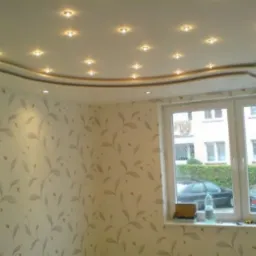 Sufit podwieszany z oświetleniem punktowym i dekoracyjną listwą, ściany pokryte tapetą w delikatny roślinny wzór, widok z okna na budynek mieszkalny.