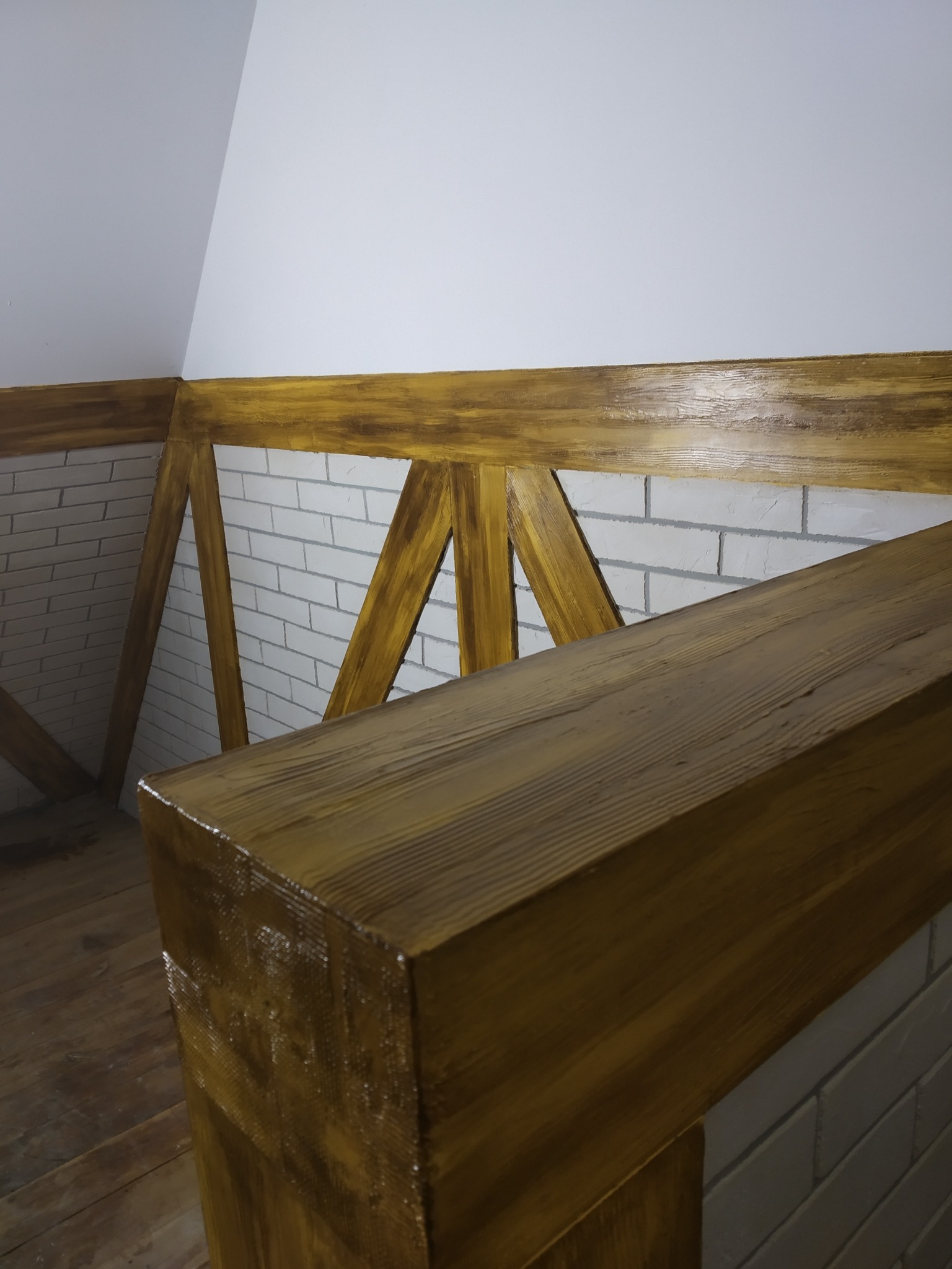 Drewniana balustrada antresoli z bejcowaną ramą i wypełnieniem w stylu rustykalnym, na tle ściany z białej cegły i drewnianej podłogi.