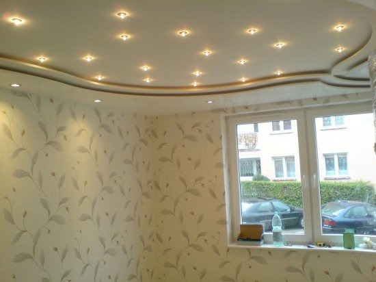 Sufit podwieszany z oświetleniem punktowym i dekoracyjną listwą, ściany pokryte tapetą w delikatny roślinny wzór, widok z okna na budynek mieszkalny.