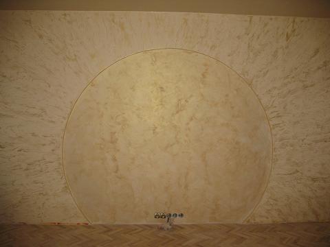 Ściana z dekoracyjnym wykończeniem w odcieniach beżu i złota, z wyraźnie zaznaczonym półokręgiem, widoczne gniazdka elektryczne przy podłodze z parkietu.