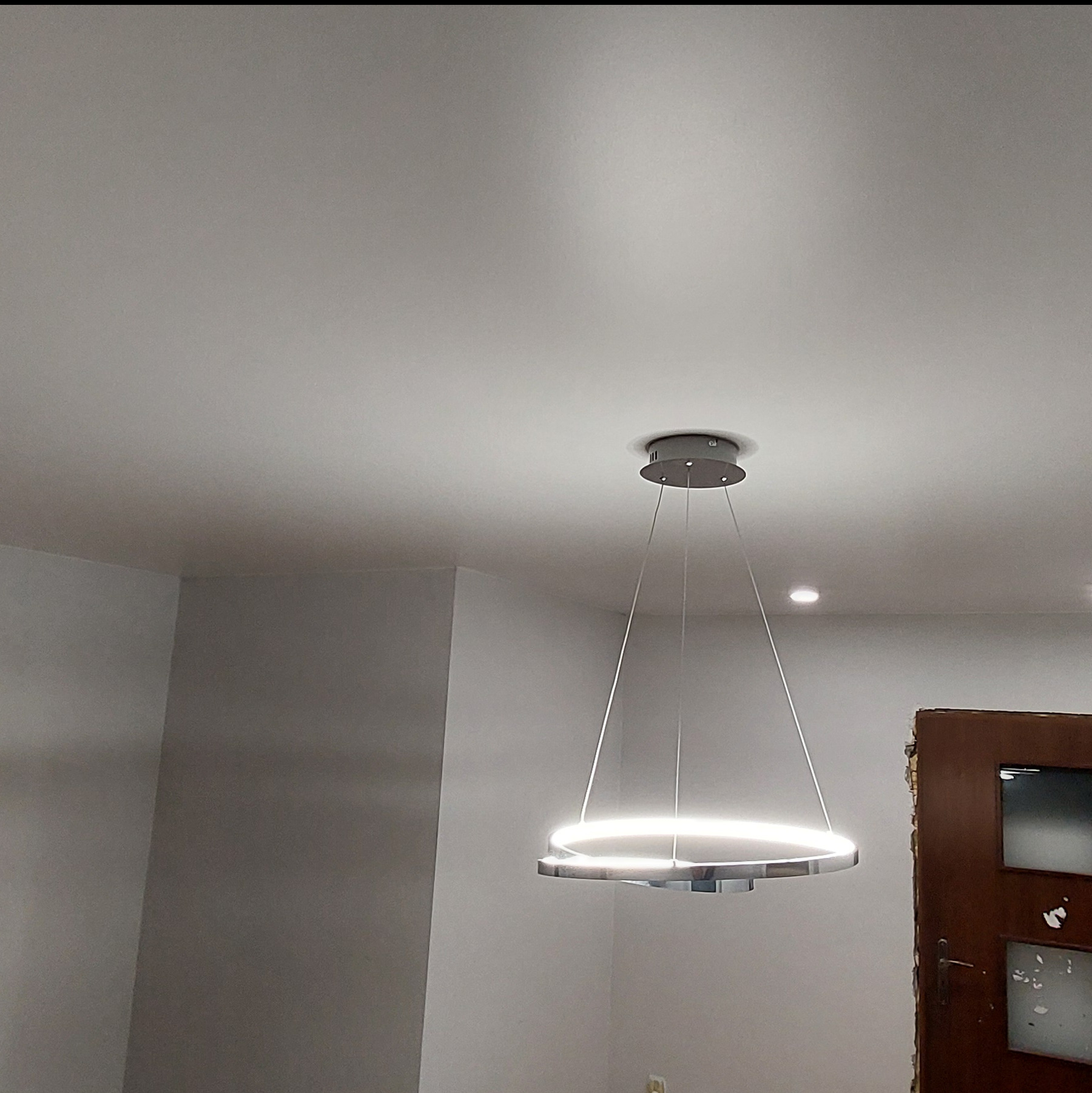 Nowoczesny, okrągły żyrandol LED zawieszony na białym suficie. Widoczne cienkie linki i minimalistyczny design lampy. W tle fragment ściany i drzwi.