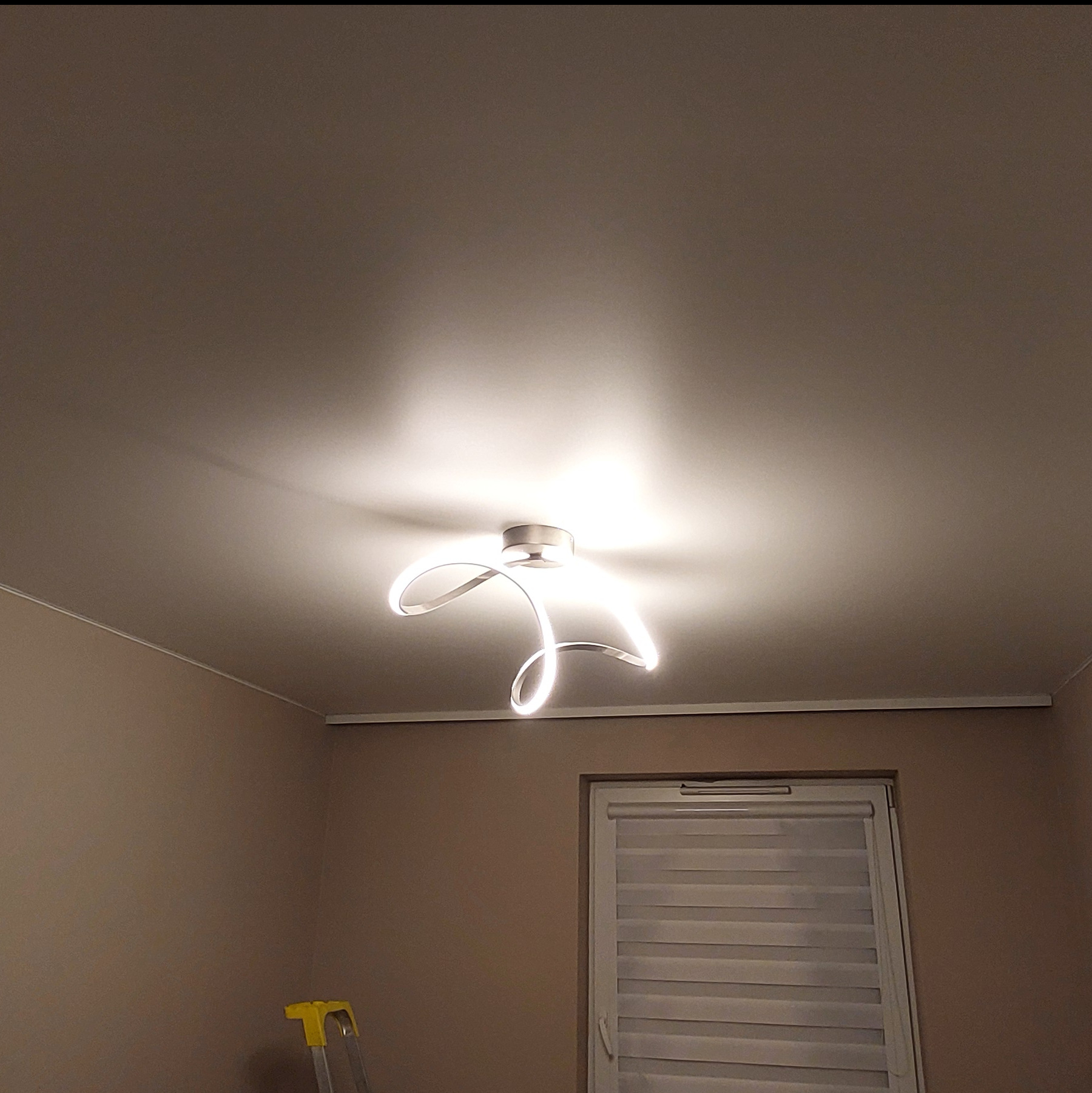 Nowoczesny, biały sufit z designerską lampą LED w kształcie wstęgi, zamontowany w pokoju w Miałach. Widoczny fragment drabiny.