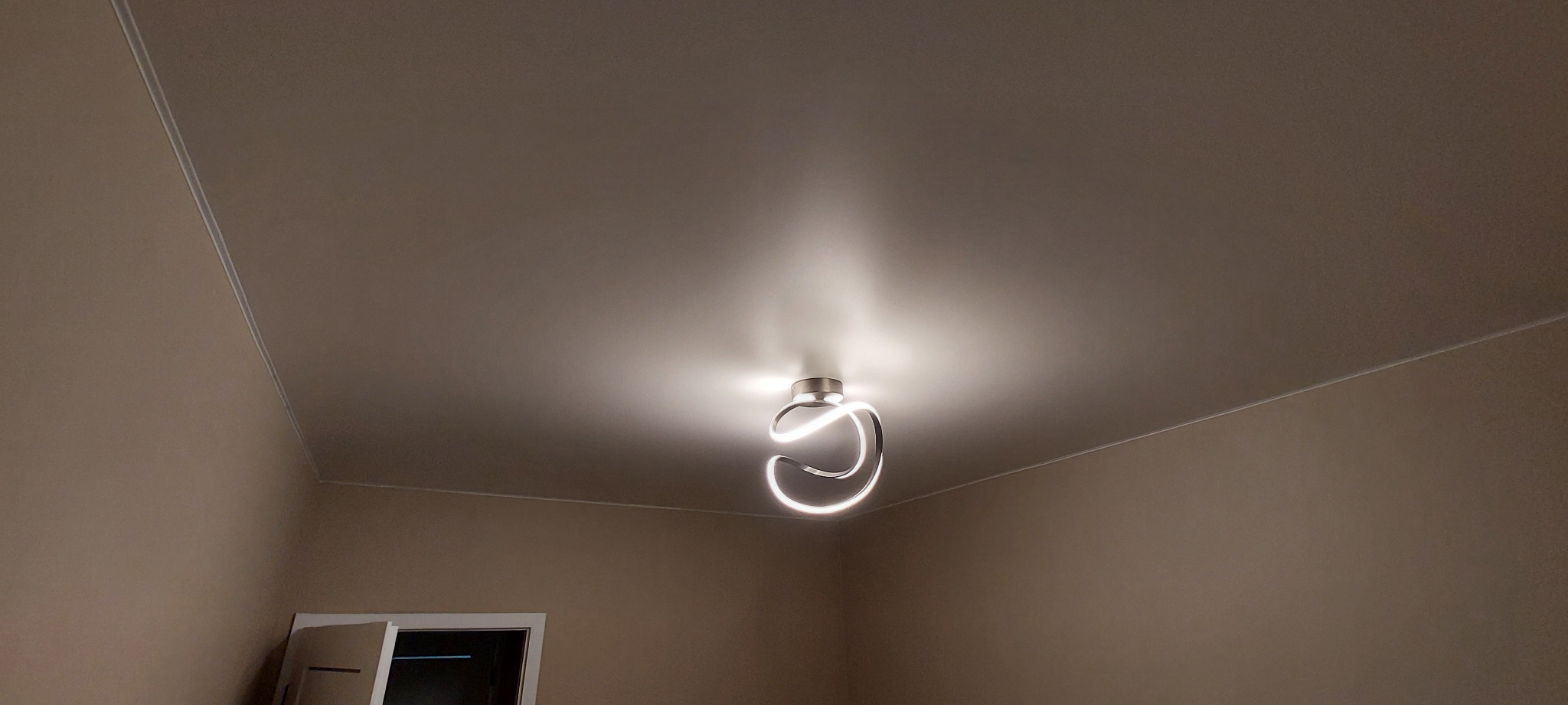 Beżowy sufit napinany z nowoczesną lampą LED w kształcie spirali, oświetlającą pomieszczenie. Widoczne listwy wykończeniowe i fragment ściany z oknem.