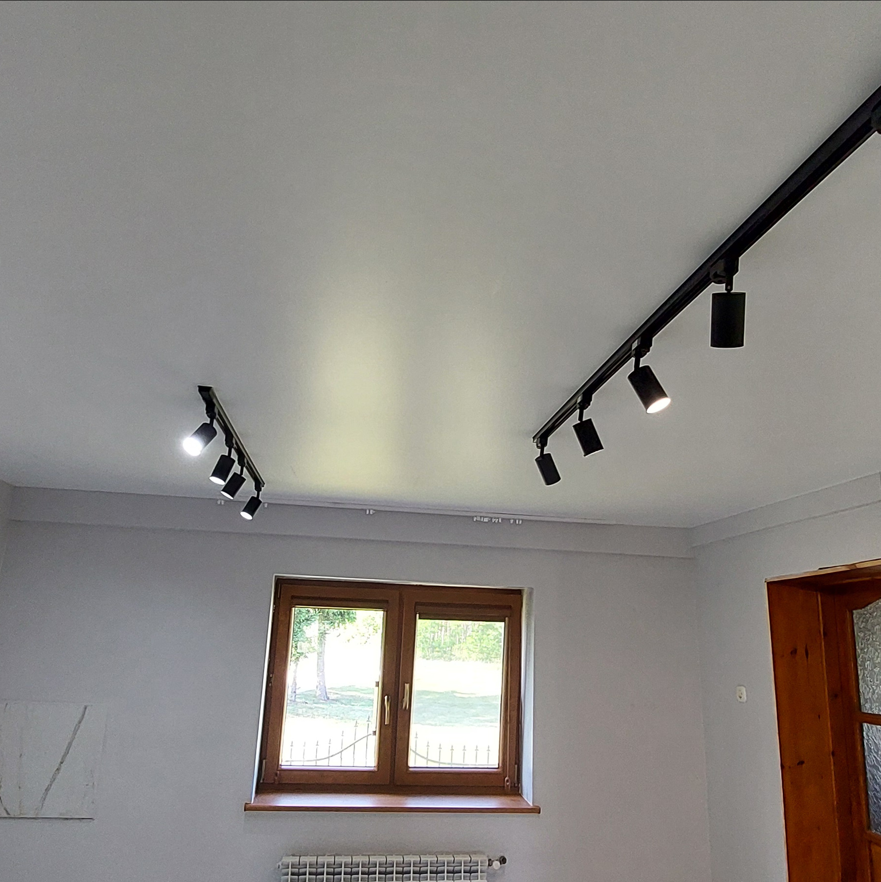 Biały sufit z zamontowanym czarnym systemem szynoprzewodów oświetleniowych z reflektorami punktowymi. Widoczne okno i fragment ściany z obrazem.