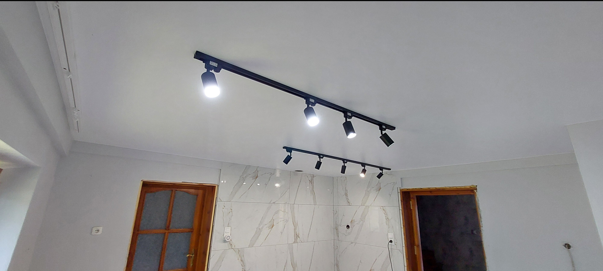 Biały sufit z dwoma czarnymi szynoprzewodami z reflektorami punktowymi. Widoczne fragmenty ścian i drzwi. Oświetlenie wnętrza.