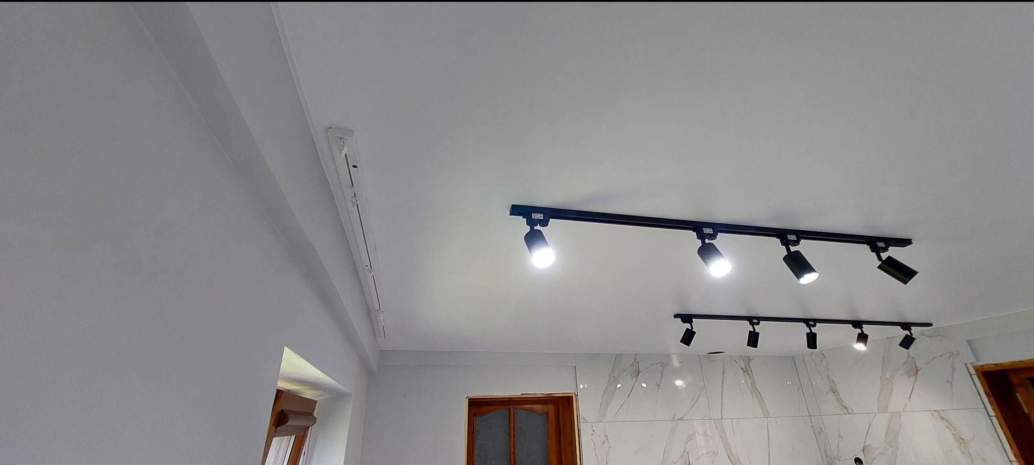 Biały sufit z zamontowanym systemem szynowym z reflektorami punktowymi. Widoczne elementy wykończenia i oświetlenie. Ujęcie z dołu, perspektywa wnętrza.