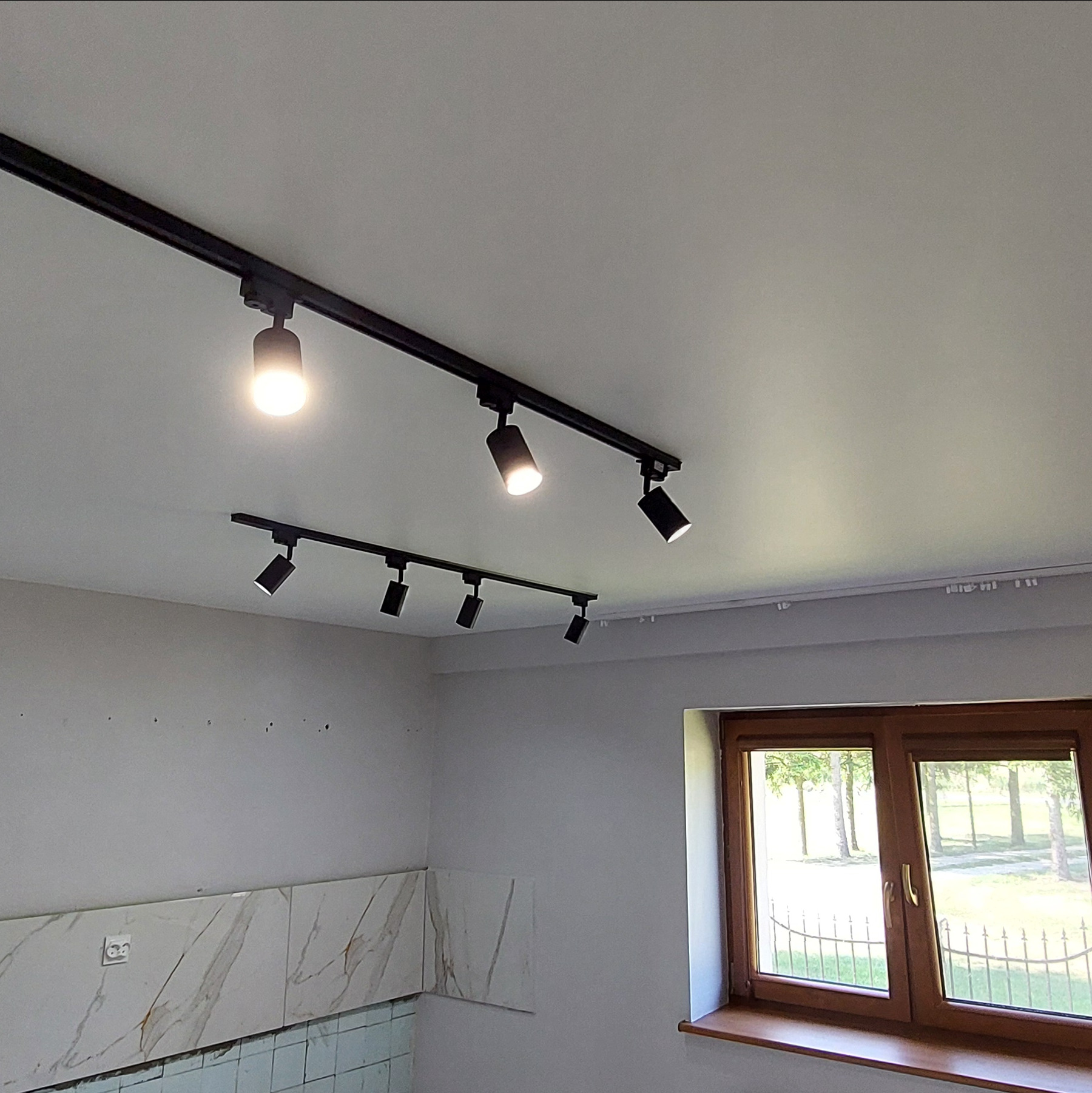 Biały sufit z dwoma czarnymi szynoprzewodami z reflektorami. Widoczne fragmenty ścian i okno z widokiem na zieleń. Oświetlenie punktowe w nowoczesnym wnętrzu.