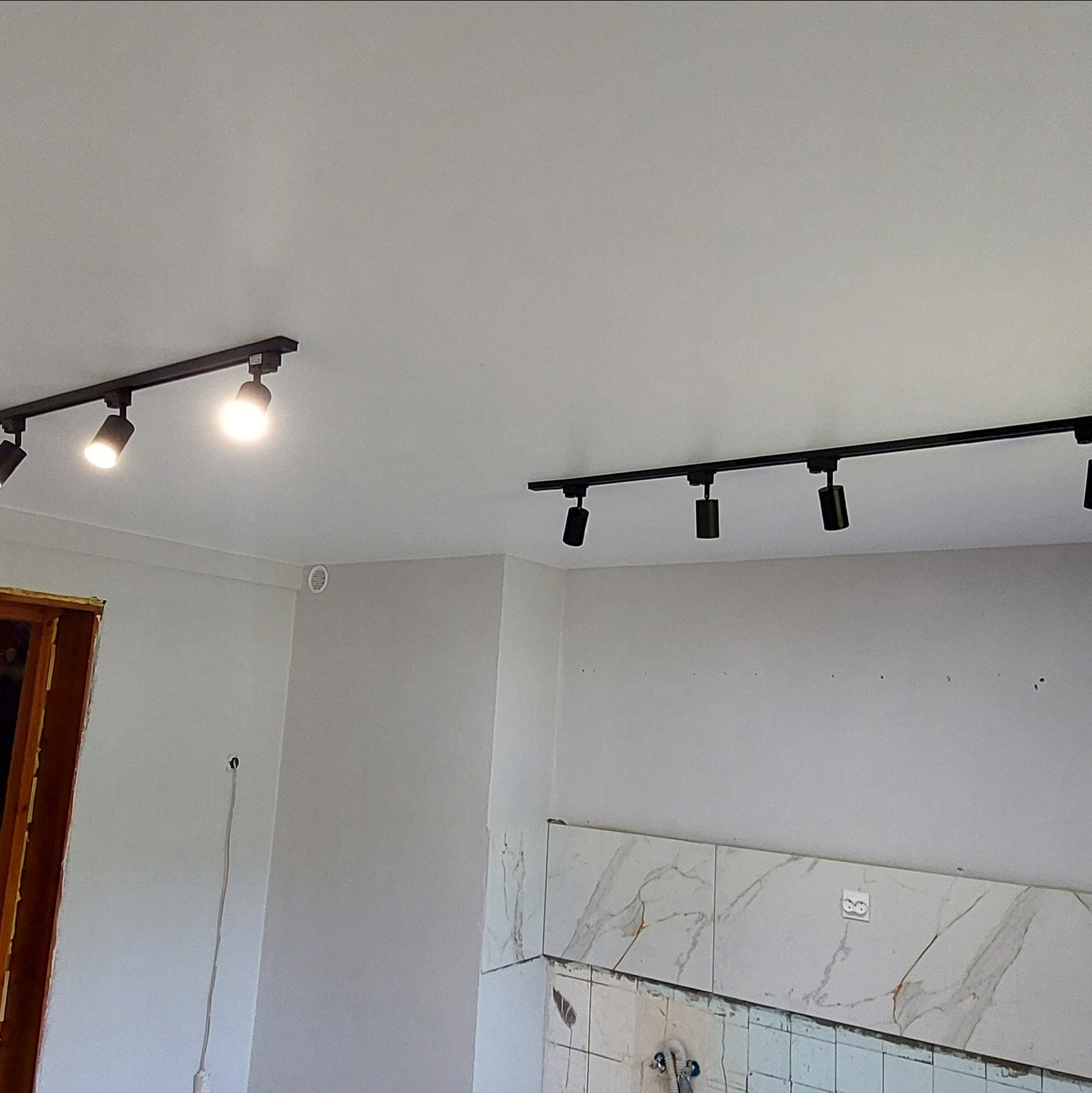 Biały sufit z zamontowanym czarnym oświetleniem szynowym z reflektorami. Widoczne fragmenty ścian z płytkami i instalacją elektryczną.