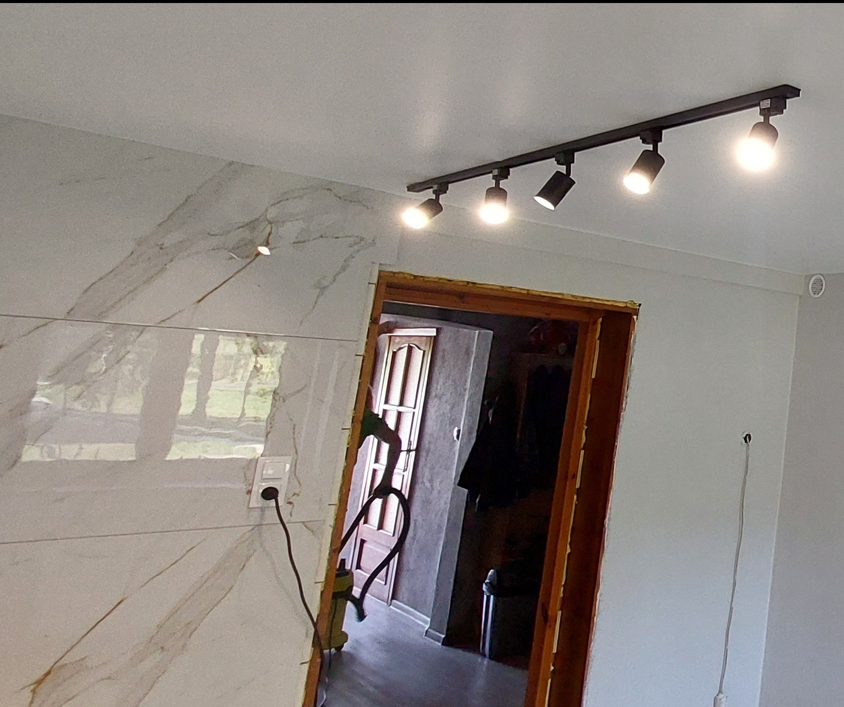Nowoczesne oświetlenie szynowe z pięcioma reflektorami, zamontowane na suficie w pomieszczeniu z marmurową ścianą i widocznym wejściem do innego pokoju.