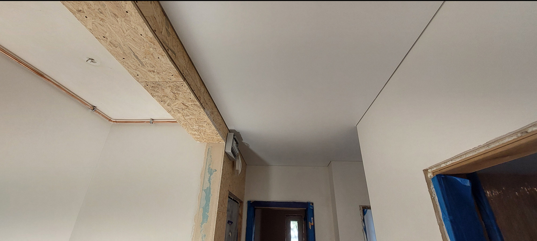 Biały sufit z widoczną konstrukcją z płyty OSB i instalacją elektryczną. Fragment wykończonego pomieszczenia z widocznymi drzwiami w trakcie zabezpieczania folią.
