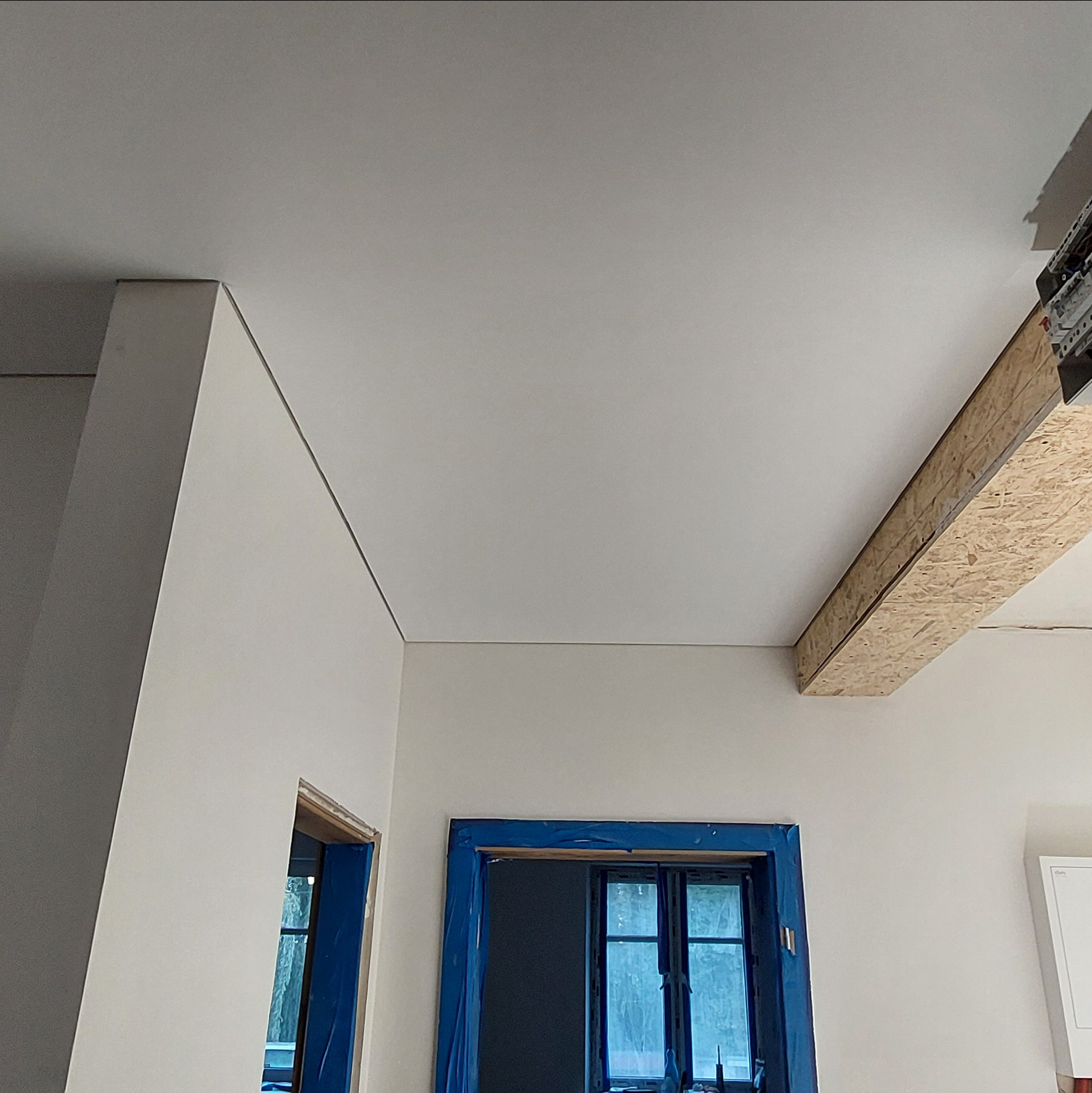 Jasne wnętrze z zamontowanym, gładkim sufitem. Widoczne zabezpieczone okna i fragment instalacji elektrycznej. Minimalistyczny design, świeżo po montażu.