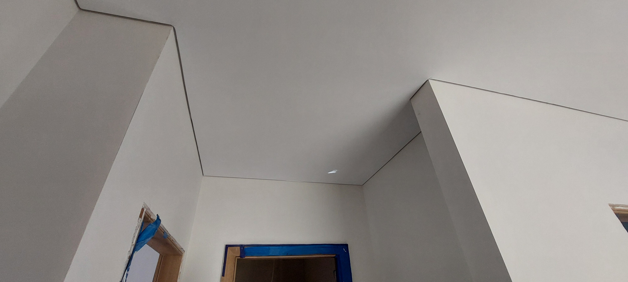 Biały, gładki sufit z delikatnym obramowaniem, zamontowany w rogu pomieszczenia. Widoczne fragmenty ścian i otwory drzwiowe zabezpieczone folią.