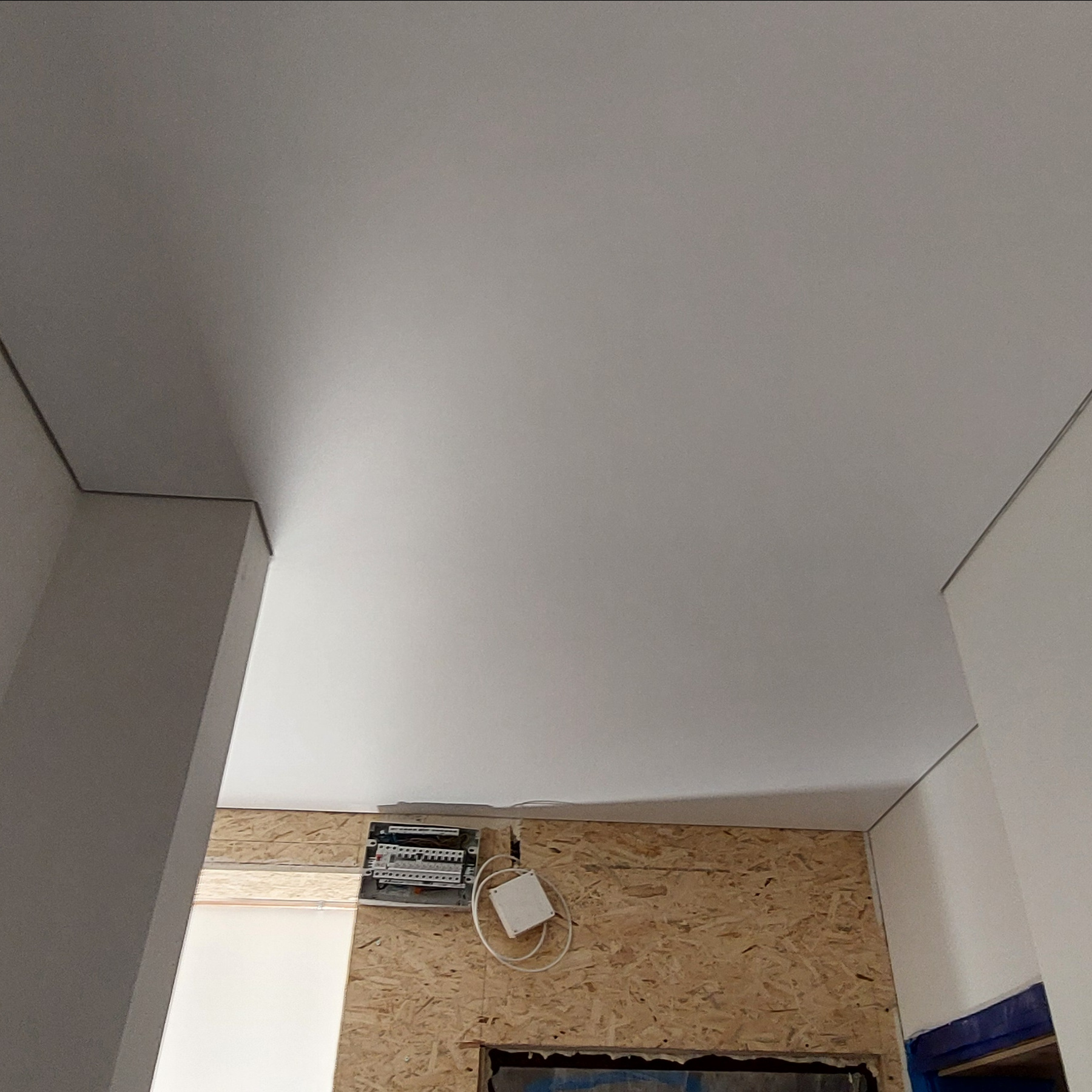 Biały, gładki sufit po montażu, widoczny z dołu, z fragmentem ściany z płyt OSB i instalacją elektryczną. Minimalistyczne wykończenie wnętrza.