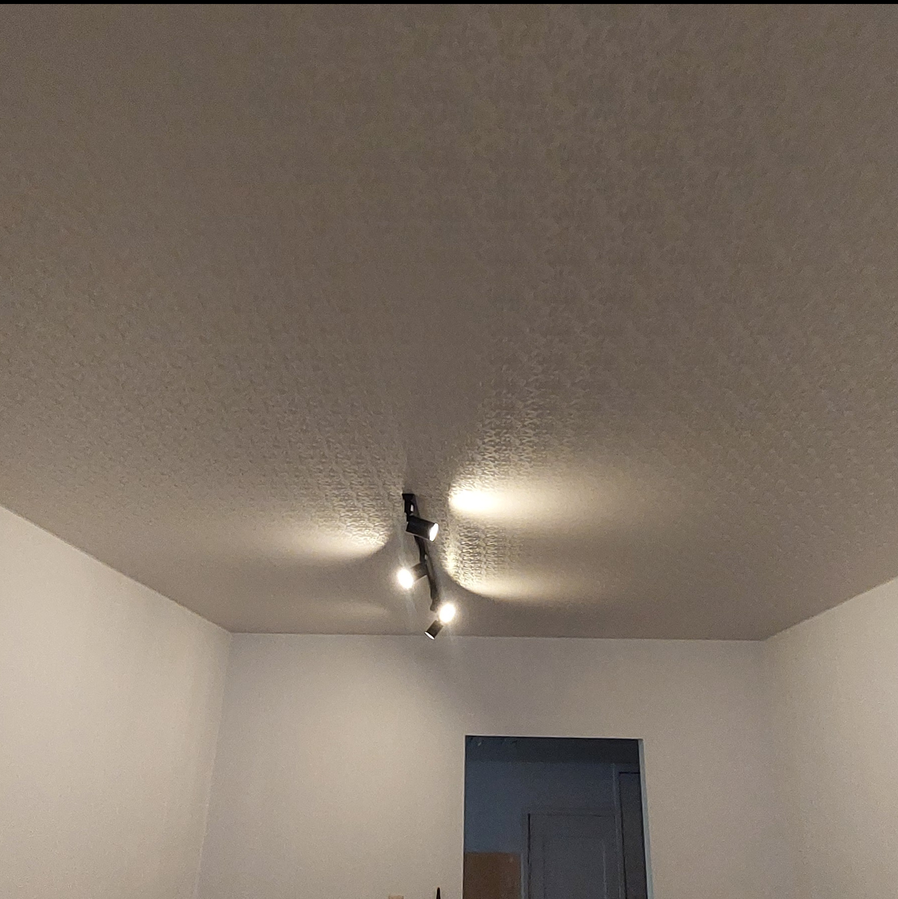 Jasny sufit napinany z delikatnym wzorem, oświetlony przez czarną, nowoczesną lampę z czterema reflektorami. Widoczne białe ściany i fragment drzwi.