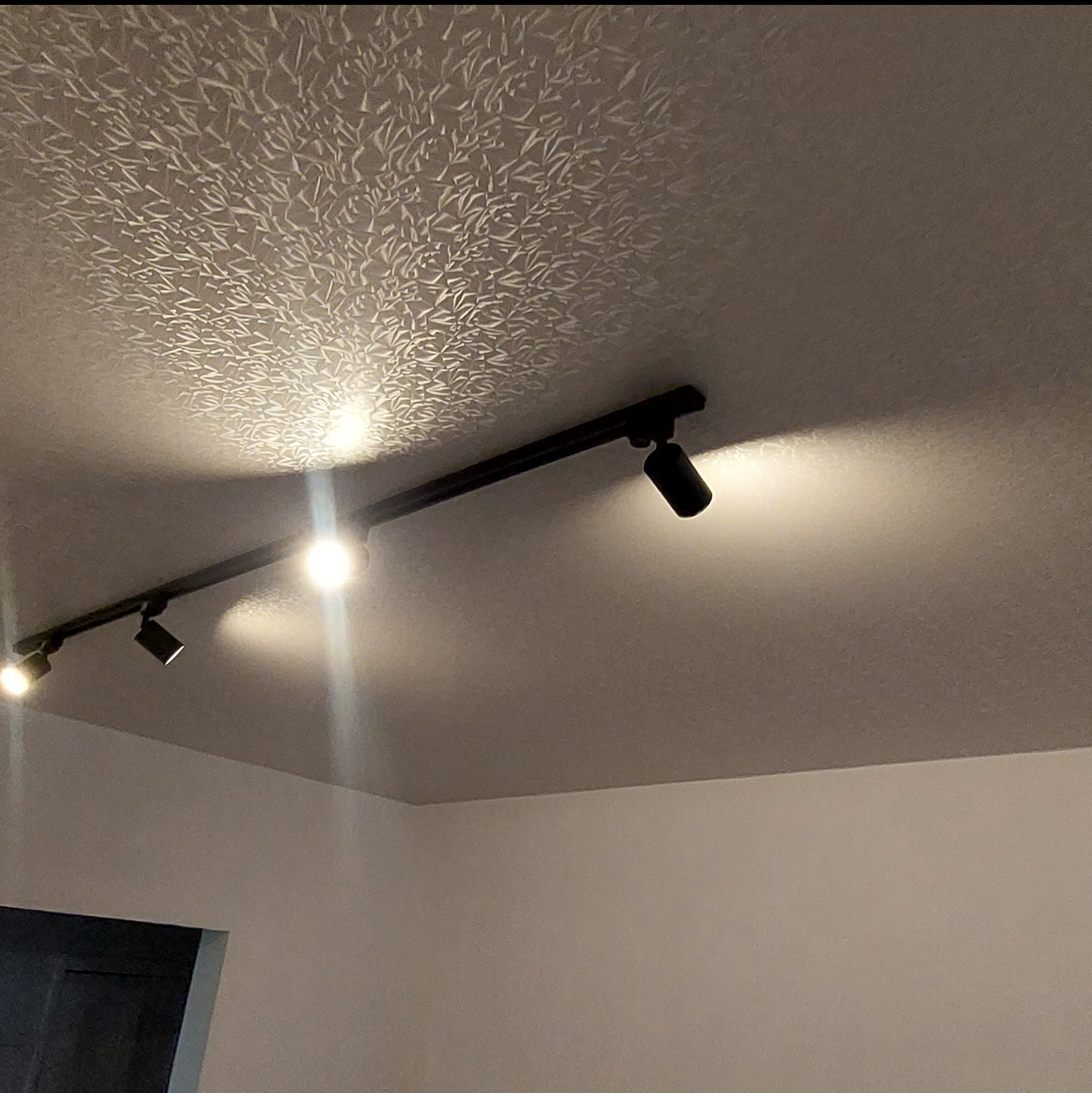 Wzorzysty sufit z zamontowanym czarnym systemem szynowym z reflektorami punktowymi, oświetlającymi powierzchnię. Dekoracyjne wykończenie sufitu.