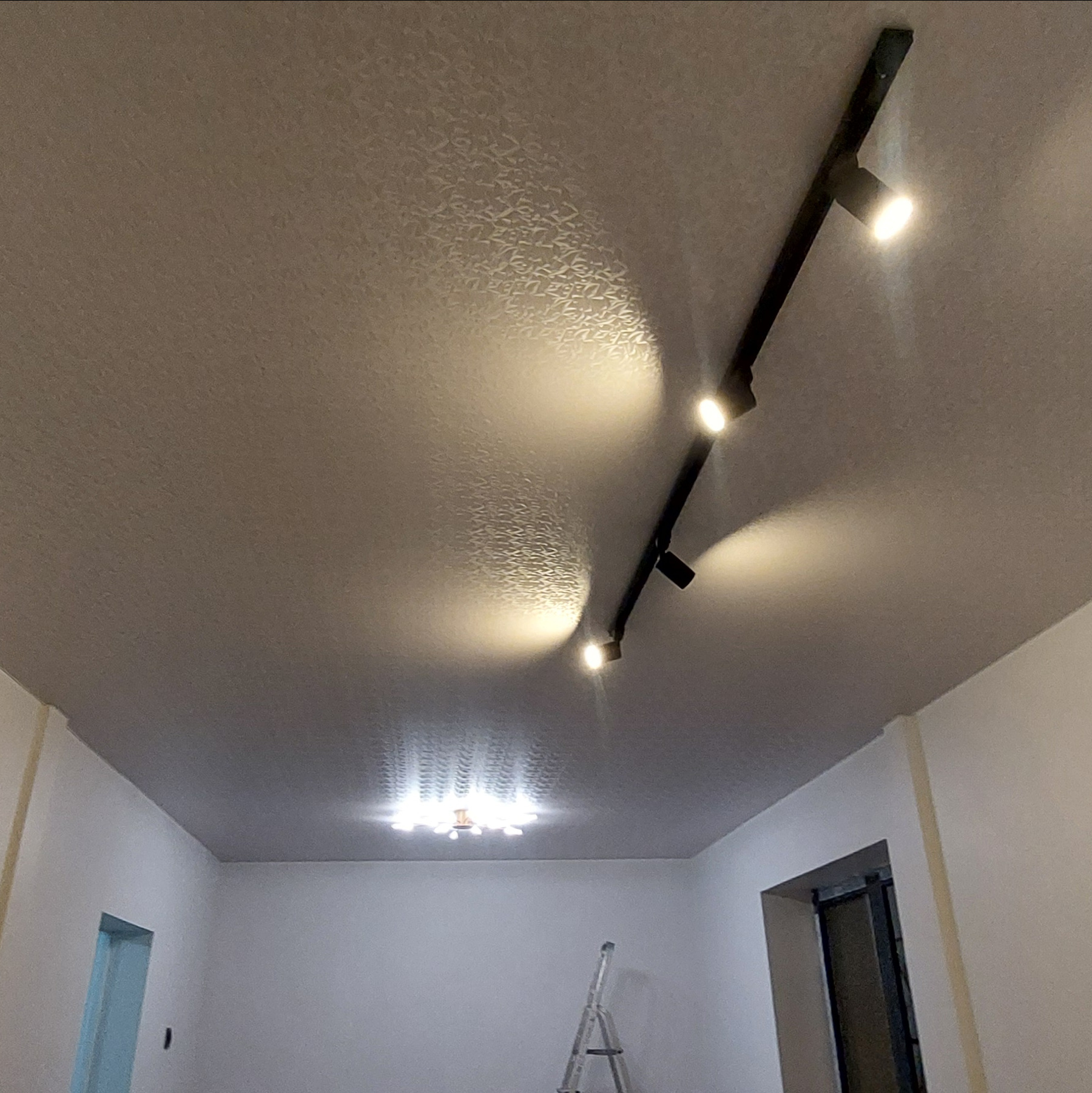 Wzorzysty sufit z zamontowaną czarną szyną oświetleniową z czterema reflektorami. W tle widoczna drabina i lampa sufitowa.