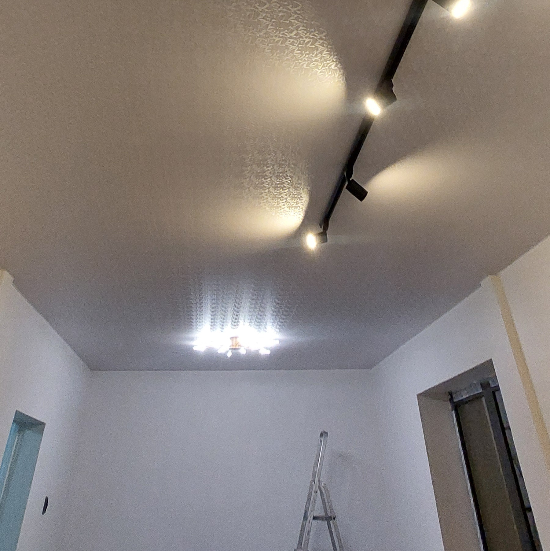 Jasny sufit z delikatnym wzorem, oświetlony reflektorami na czarnej szynie i centralnym żyrandolem. W tle ściana i drabina malarska.