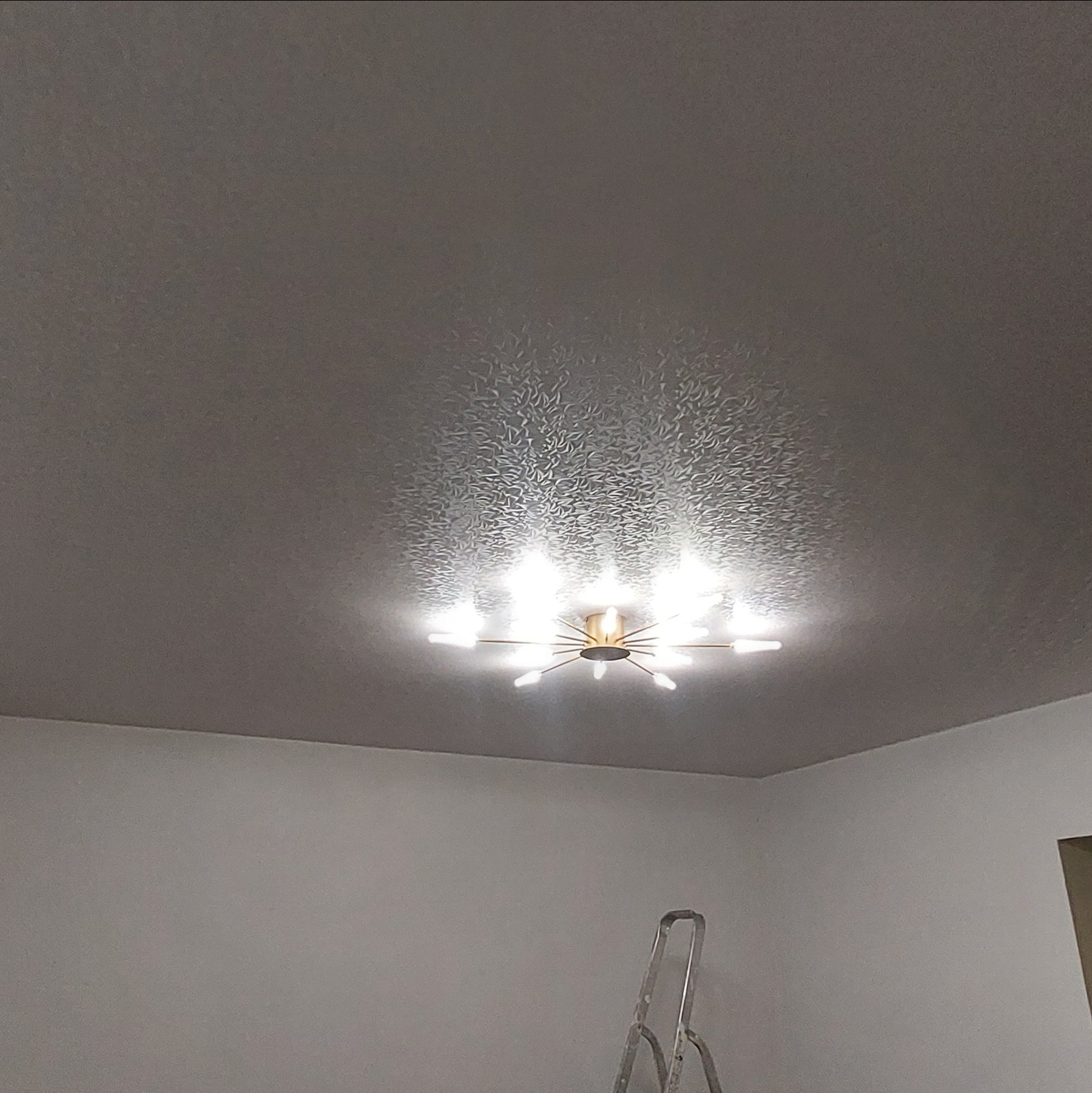 Jasnoszary sufit z delikatnym wzorem i nowoczesną lampą. W rogu widoczna drabina, sugerująca prace wykończeniowe.