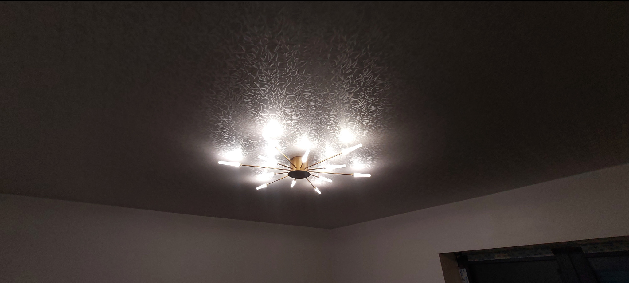 Sufit napinany z wzorem i nowoczesną lampą w stylu gwiazdy, oświetlającą pomieszczenie. Widok z dołu, podkreślający estetykę wykończenia.