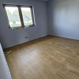 Filip Grzywacz - Pokój po remoncie: świeżo pomalowane szare ściany, ułożona podłoga z paneli imitujących drewno. Widoczne okno i wystające kable elektryczne.