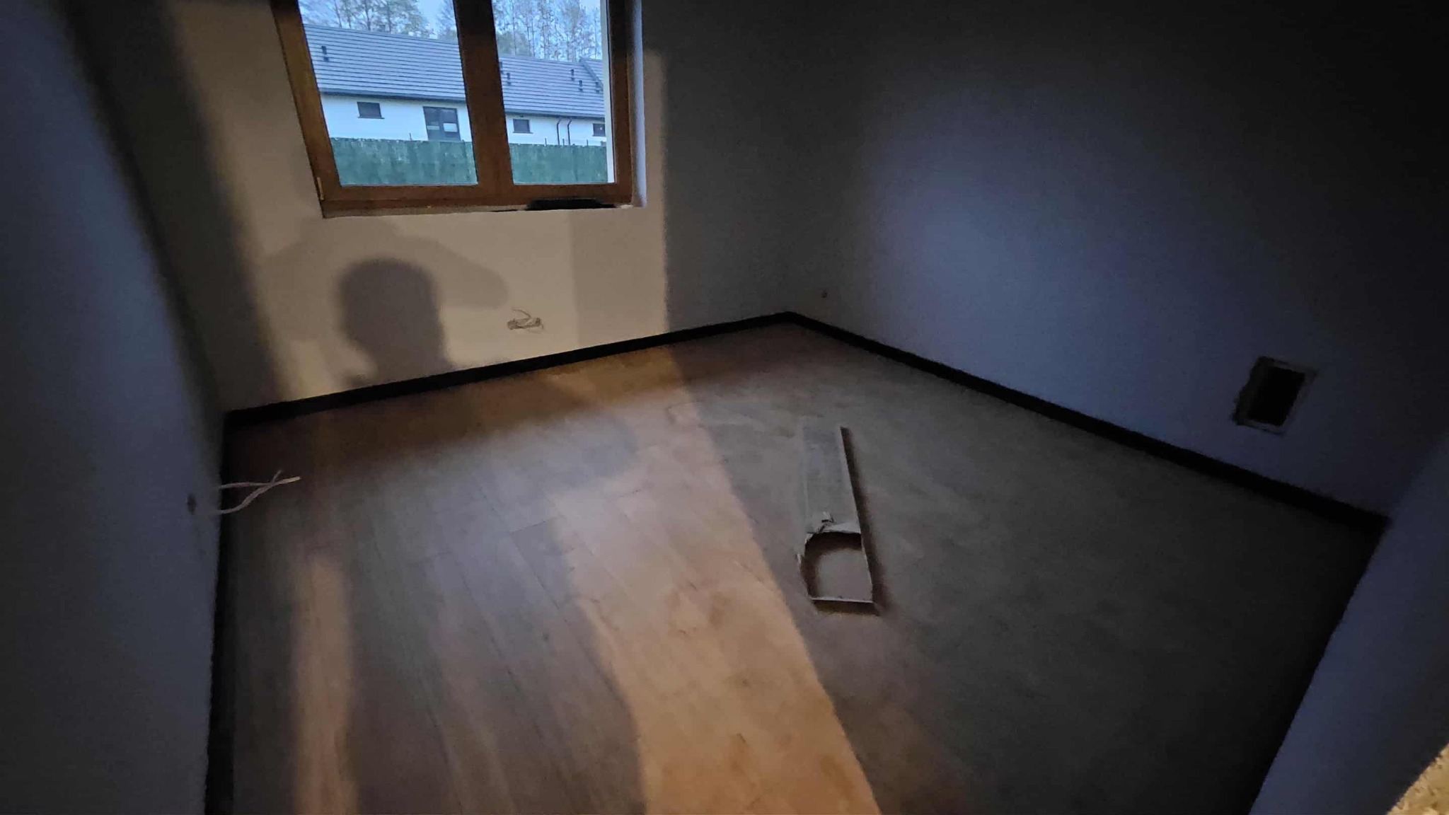 Jasne panele podłogowe w pokoju z oknem, widoczne kable przy ścianie i fragment kartonu na podłodze. Ujęcie z rogu pomieszczenia, w tle widok na dom.