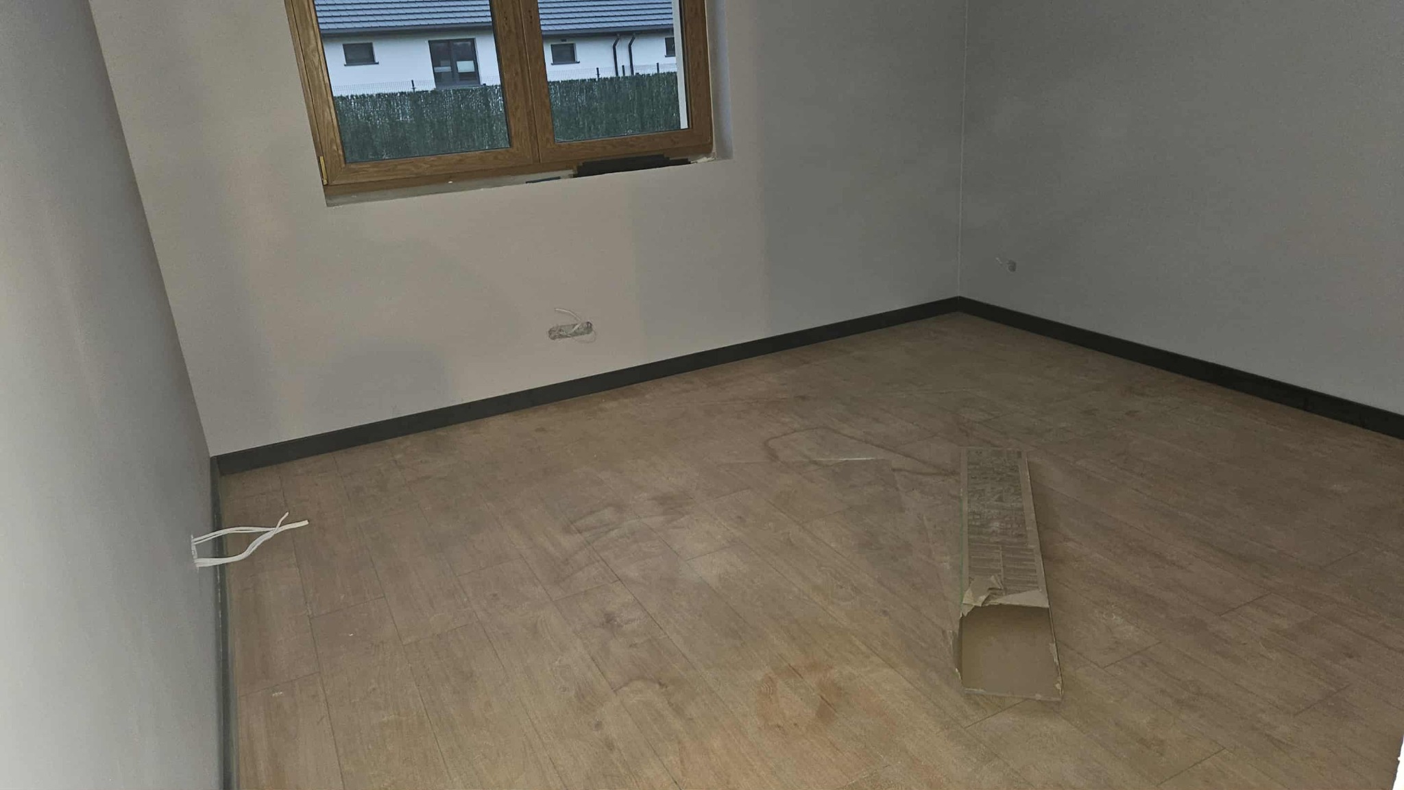Ułożone panele podłogowe w pokoju z oknem, listwy przypodłogowe w kolorze grafitowym. Widok na podłogę w Łodzi. Częściowo widoczny karton po panelach.