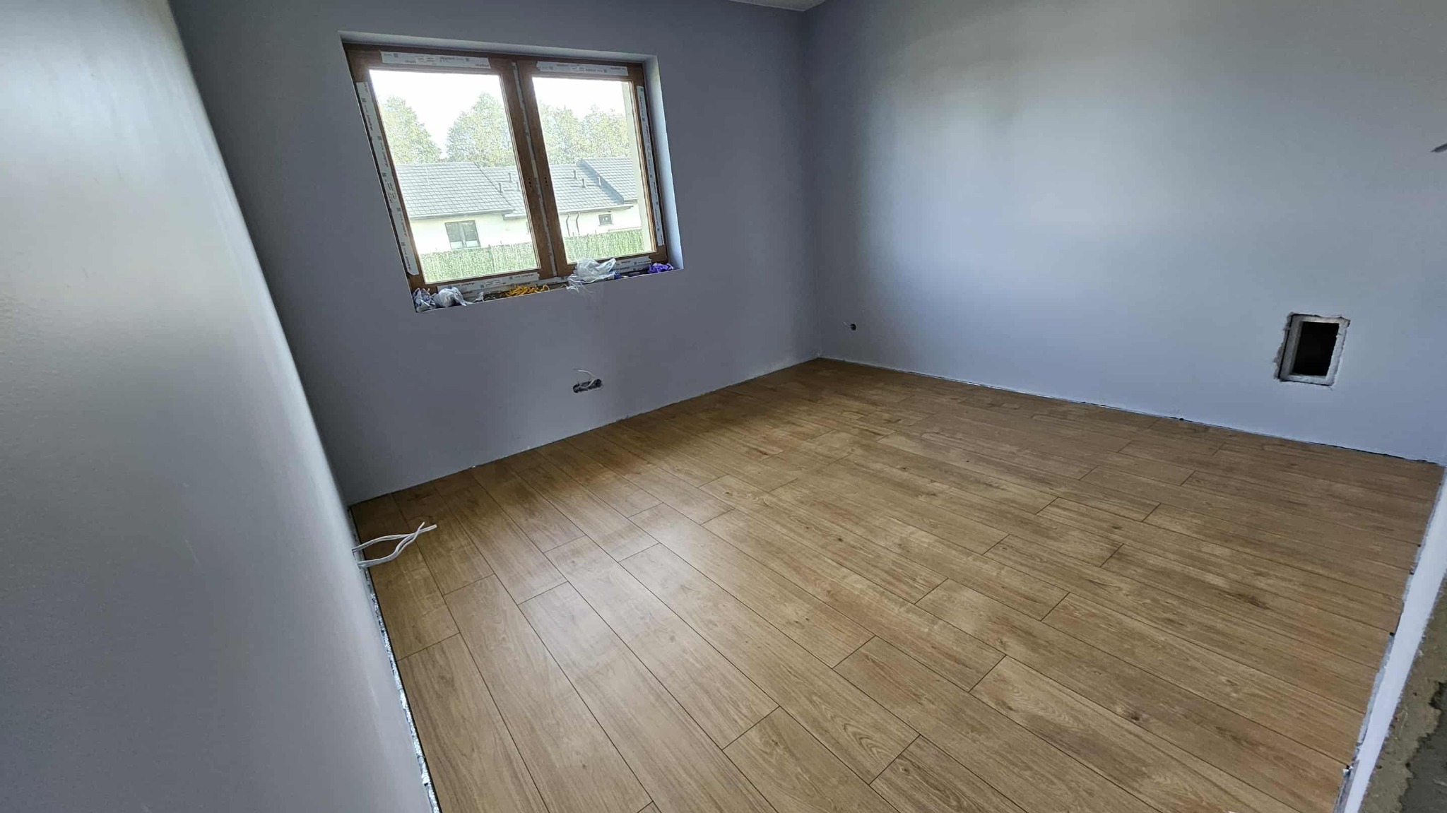 Pokój po remoncie: świeżo pomalowane szare ściany, ułożona podłoga z paneli imitujących drewno. Widoczne okno i wystające kable elektryczne.