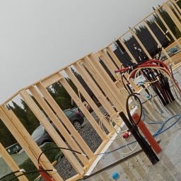 Filip Grabiński Nowoczesne Budownictwo - Wznoszona konstrukcja domu szkieletowego z widocznymi elementami instalacji elektrycznej i hydraulicznej, na zewnątrz widoczny samochód osobowy i drzewa.