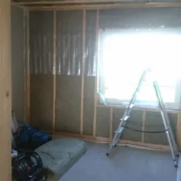 Wnętrze w trakcie ocieplania: wełna mineralna między drewnianymi belkami na ścianach i suficie, folia paroizolacyjna, drabina aluminiowa przy oknie, kompresor Wurth na podłodze.