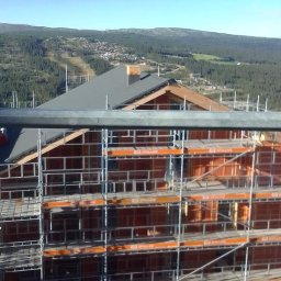 Filip Grabiński Nowoczesne Budownictwo - Wznoszony dom z drewnianą konstrukcją szkieletową, widoczny z rusztowania na tle zalesionych wzgórz i błękitnego nieba.