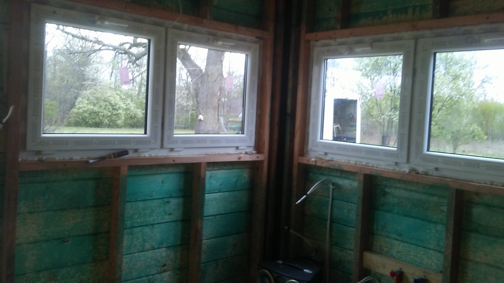 Wnętrze w trakcie budowy z dwoma zamontowanymi oknami PCV, widoczne zielone deski ścian i drewniana konstrukcja szkieletowa.