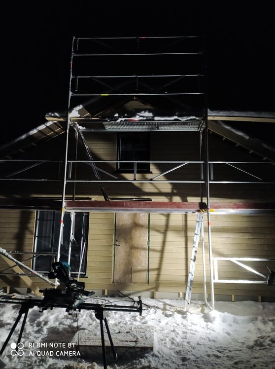 Montaż rusztowania przy ocieplonym poddaszu drewnianego domu w zimowej scenerii, widoczna piła ukośnica.