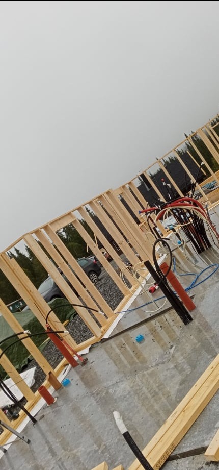 Wznoszona konstrukcja domu szkieletowego z widocznymi elementami instalacji elektrycznej i hydraulicznej, na zewnątrz widoczny samochód osobowy i drzewa.