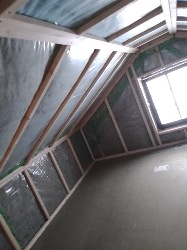 Wnętrze poddasza z drewnianą konstrukcją szkieletową, folią izolacyjną i wełną mineralną, widoczne okno dachowe i wstępnie otynkowana ściana.
