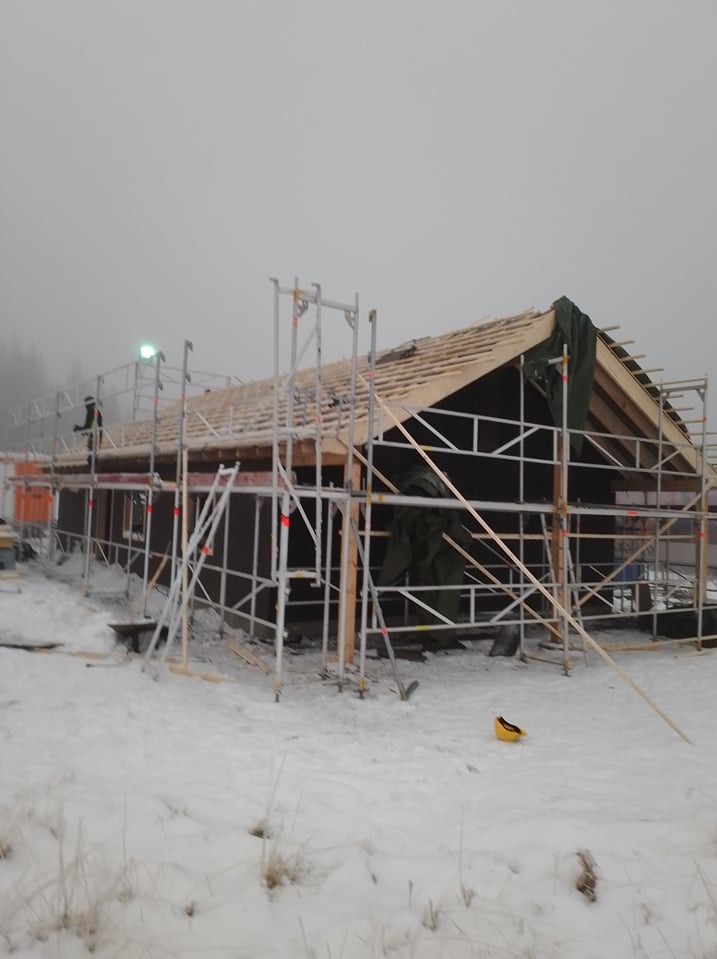 Budowa domu w zimowej scenerii; widoczne rusztowania, drewniana konstrukcja dachu, czarne panele ścienne i pracownik w oddali.