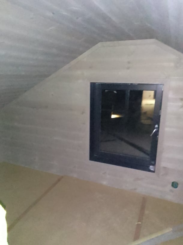 Wnętrze poddasza z drewnianą boazerią i oknem w czarnej ramie, widoczne niedoskonałości wykończenia i fragment podłogi z płyt OSB.