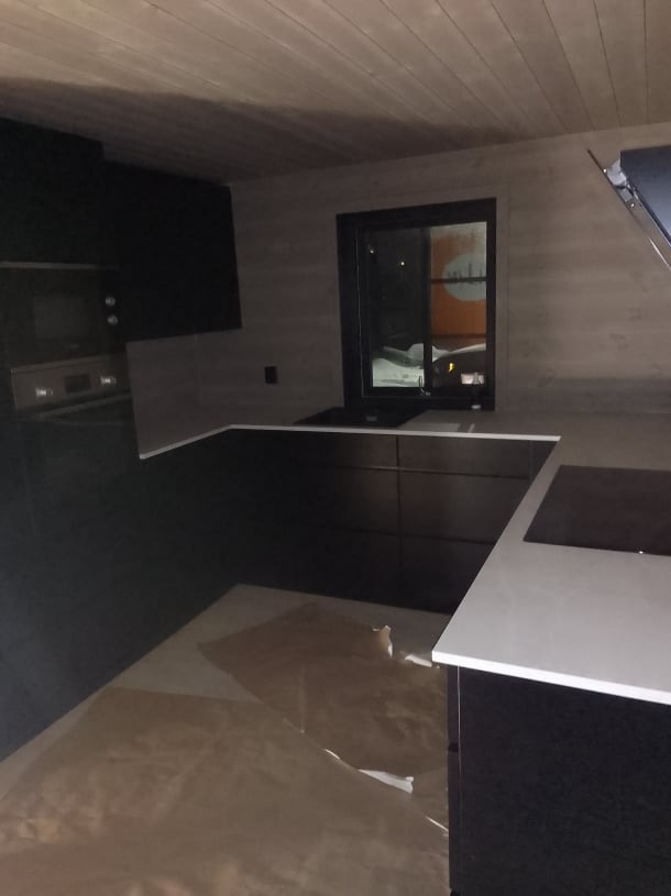 Nowoczesna, czarna zabudowa kuchenna z białym blatem w domu z drewnianym sufitem, widok na okno.