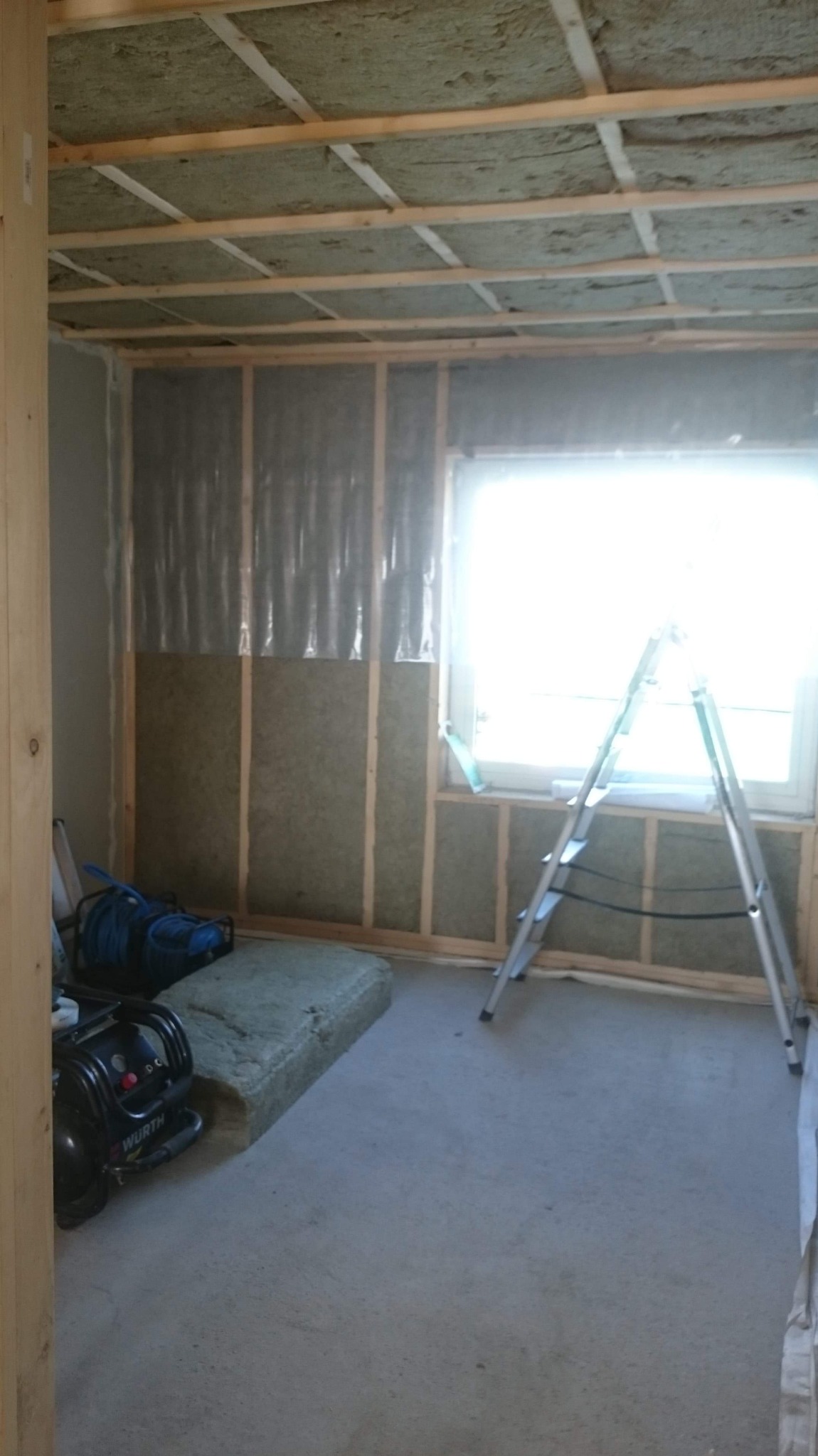 Wnętrze w trakcie ocieplania: wełna mineralna między drewnianymi belkami na ścianach i suficie, folia paroizolacyjna, drabina aluminiowa przy oknie, kompresor Wurth na podłodze.